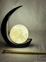 Lamp lit, showing glowing moon globe within black crescent base, measuring tape for scale