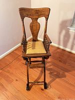 Front view of wooden antique baby high chair showing spindled arms, seat cushion, footrest and wheels