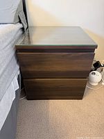 Nightstand beside bed showing dark brown finish, two drawers, glass top