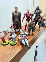 Group of eight figures on wooden rail including Iron Man, brown-gold armoured male, muscular warrior, hooded assassin, Lock, Shock, Barrel and Sally