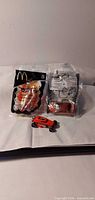Two sealed McDonald’s Hot Wheels bags and loose orange car on surface