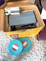 Projector in original box with film reels and splicing tape visible