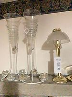 Pair of tall glass hurricane candle holders on white metal stands beside small brass lamp