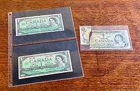 Two 1967 $1 banknotes in protective sleeve beside torn 1973 note front side
