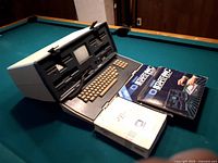 Osborne computer open on table showing screen, drives, keyboard, plus three manuals beside it