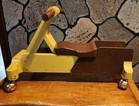 Side view of wooden ride-on scooter showing yellow frame, brown seat, handle, and ball casters