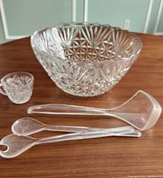 Clear pressed-glass bowl, one glass cup, and three clear plastic serving utensils on wooden surface