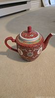 Left side view of red porcelain teapot showing spout, handle, floral design, Chinese characters