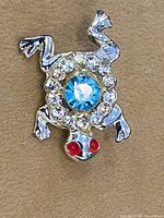 Front view showing frog shape, blue center crystal, clear rhinestones, red eyes