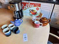 Entire lot showing coffee maker, glass carafe, both teapots, mugs, creamer, and metal coffee sign