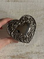Hand holding heart-shaped silver-plated lid showing ornate floral scroll design