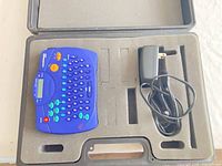 Blue DYMO handheld label maker in foam insert with AC adapter inside black carrying case