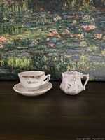Teacup on saucer beside ornate creamer in front of painting backdrop
