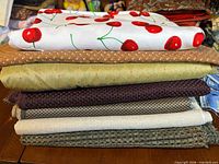 Stack of seven folded fabric pieces showing cherry print, brown floral, olive-gold satin, plum woven, small check upholstery, beige linen, green tweed