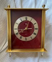 Front view of gold-tone quartz tabletop clock showing red face and Roman numeral dial