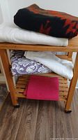 Stack of four visible blankets on wooden shelf: black/red, white, purple floral, bright pink
