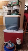 Overall lot showing two coolers stacked, lantern, boxed lamp, beverage jug, plates, glove, can opener