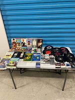 Overall lot on table showing vinyl singles, cassettes, VHS tapes and DVDs