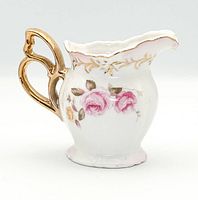 Side view of Lefton porcelain creamer showing pink rose sprays, gilt ornate handle, scalloped rim, footed base