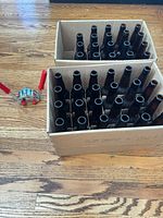 Two boxes containing empty dark amber long-neck beer bottles; red-handled crown capper beside boxes