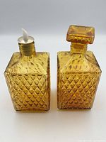 Front view of two amber square pressed glass decanters, showing diamond pattern, original stopper on right bottle and replacement pour spout on left bottle