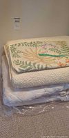 Stack of botanical bedspread atop two off-white quilted bedspreads on plastic sheet