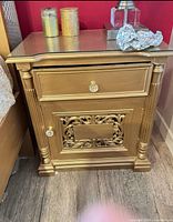 Front view of one gold wooden nightstand showing glass top, drawer, carved door, knobs, turned columns