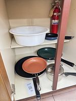 Cabinet shelves holding white basin, fire extinguisher, several frying pans, saucepan