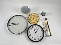 Group shot of all six items: two wall clocks, sunrise alarm lamp, cube clock, round thermometer, packaged thermometer