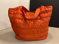 Front view of orange quilted tote showing size, color, exterior zip pocket, logo plate