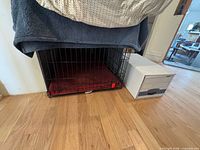 Front view of large black wire dog crate with red cushion, white storage box beside for scale