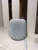 Front/side view of white Apple HomePod showing mesh exterior and attached power cord