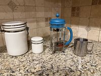 All four items lined up on countertop: large and small white clamp-lid canisters, blue-handled Bodum French press, stainless milk pitcher