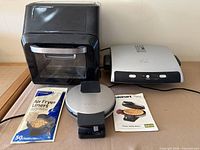 Air fryer, George Foreman grill, Cuisinart waffle maker, liners pack and manual on surface