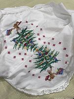 Close-up of two cross-stitched Christmas trees with gifts and toy figure on tablecloth