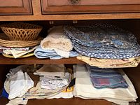 Shelf with folded blue floral quilted placemats, cream fringed runner, blue tablecloth and wicker basket