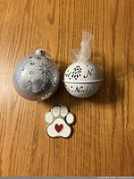 Two spherical Christmas ornaments and one paw print ornament displayed on wooden surface