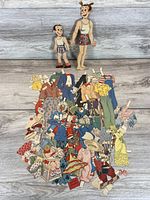 Two paper doll figures beside pile of clothing cut-outs