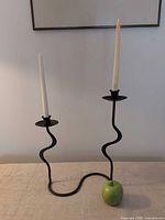 Front view of black serpentine double-arm wrought iron candleholder with two taper sockets, apple shown for scale