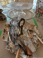 Overview showing stacked glass cake stands, silverplate utensils and porcelain pickle dish