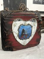 Angled view showing front of plaque, cracked mirror heart frame, central mermaid skeleton image, wire hanger