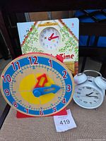 Group shot of wooden learning clock, Usborne board book, white twin-bell alarm clock, lot tag visible