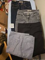 Four pairs of pants stacked: dark indigo jeans, gray jeans, black pants, light blue pants