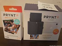 Front view of boxed Prynt Pocket printer and boxed 60-pack Zink paper