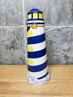 Front view of ceramic lighthouse coin bank showing white body, blue spiral stripes, yellow windows, molded base