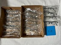 Two cardboard trays containing rows of individually bagged eyeglasses plus additional pairs to the side