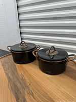 Two Neva black casserole pots with lids on wooden surface
