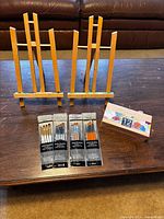 Two small wooden easels standing, four sealed brush packs, one sealed 12-colour acrylic paint set displayed on tabletop
