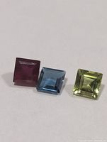 Three square-cut gemstones aligned—ruby left, blue topaz center, peridot right