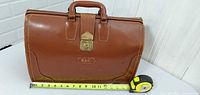 Front view of brown leather doctor’s bag with handle, brass lock, tape measure showing width ~11–12 in